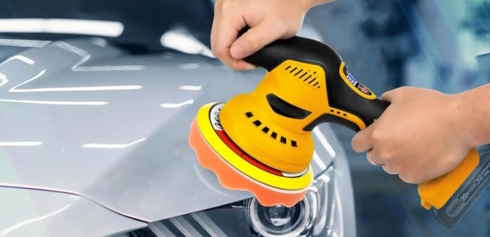 Close-up of a person using a yellow cordless buffer to polish a silver car hood.
